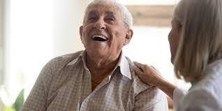 An elderly man laughs as a woman rests her hand on his shoulder.